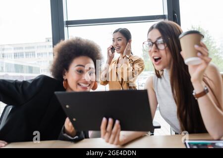 des responsables interraciaux enthousiastes regardant le presse-papiers à proximité tandis que la femme d'affaires asiatique heureuse parle sur un smartphone Banque D'Images