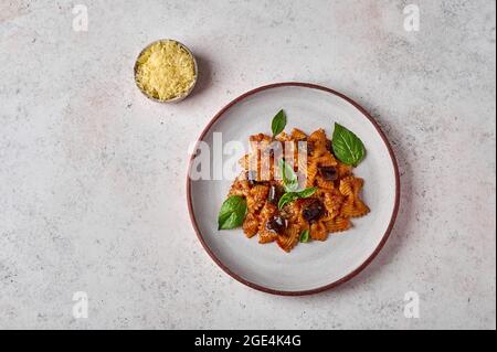 Pâtes farfalle à l'aubergine, sauce tomate, fromage dans un bol et basilic sur une assiette sur fond texturé gris, vue du dessus, espace copie Banque D'Images