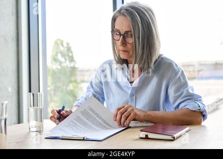 Femme d'affaires sérieuse senior en lunettes de lecture contrat de partenariat à la table. Banque D'Images