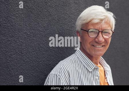 Portrait de la tête et des épaules d'homme moderne senior regardant l'appareil photo et souriant en se tenant debout près du mur à l'extérieur, espace de copie Banque D'Images