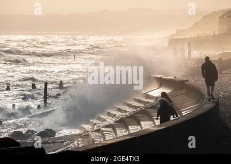 Les gens brave les vagues féroces qui s'écrasant sur les défenses de la mer à Milford-on-Sea dans le Hampshire en été avec des falaises lointaines en plein soleil. Banque D'Images