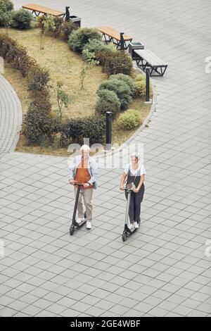 Vue verticale en grand angle sur un couple contemporain, à cheval sur des scooters électriques dans un parc sur fond carrelé, espace de copie Banque D'Images