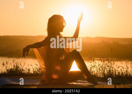 Silhouette de jeune femme assise contre le soleil dans la lumière chaude du soleil du matin, méditation yoga exercice de forme physique à l'extérieur Banque D'Images