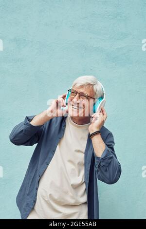 Portrait vertical de l'homme moderne senior écoutant de la musique et souriant tout en portant de grands écouteurs sur fond bleu Banque D'Images