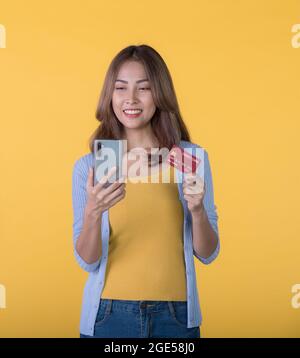 Femme asiatique enthousiaste tenant une carte de crédit et un smartphone se sentant excitée tout en regardant le téléphone mobile isolé sur fond de couleur Banque D'Images