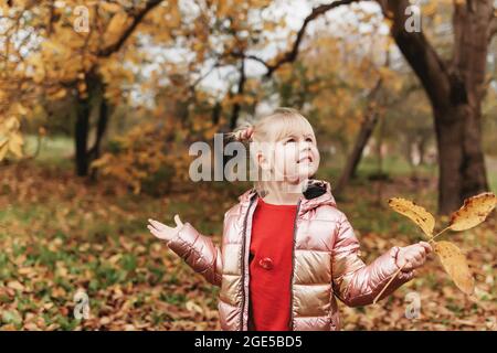 Petite fille blonde joue avec des feuilles d'automne jaunes dans le jardin, sourire, a du plaisir Banque D'Images