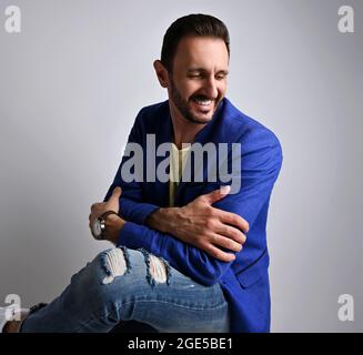 Portrait d'un homme adulte élégant en riant dans un Jean moderne déchiré et une veste bleue assis sur un tabouret sur le côté Banque D'Images