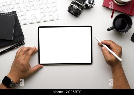 Gros plan, vue de dessus, espace de travail moderne de l'éditeur de photos avec maquette d'écran vide de tablette graphique, stylet à main levée, appareil photo, café, livres, clavier, blanc Banque D'Images