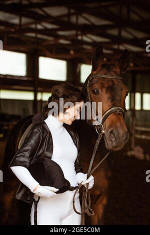 Mignonne jockey fille, à côté de beau cheval brun tir gros plan. Belle jeune fille se tient à côté de cheval dans stable. Elle porte une belle robe blanche. Ranch. Équitation. Hippodrome Banque D'Images