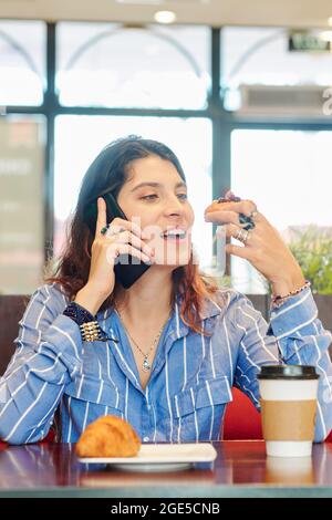 Belle jeune femme en train de savourer un délicieux dessert et de parler au téléphone lorsqu'elle est assise à une table de café Banque D'Images