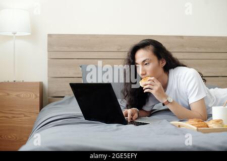 Beau jeune homme avec de longs cheveux allongé sur le lit, manger un petit pain frais et regarder un séminaire en ligne ou un tutoriel sur un ordinateur portable Banque D'Images