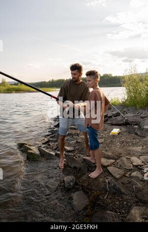 Jeune homme tournant la roue sur la tige tout en pêchant avec son fils adolescent Banque D'Images