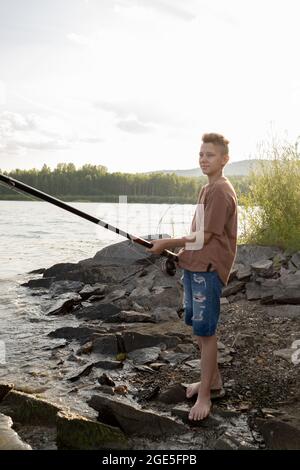 Adolescent actif avec long bâton debout au bord de l'eau tout en pêchant par lui-même Banque D'Images