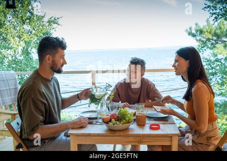 Une famille heureuse assise près d'une table servie lors d'un dîner à l'extérieur ou d'un petit déjeuner au bord de l'eau Banque D'Images