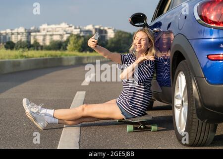 Une femme gaie prend une photo de selfie assis à bord d'une voiture moderne, admirez le coucher du soleil pendant un seul voyage en voiture Banque D'Images