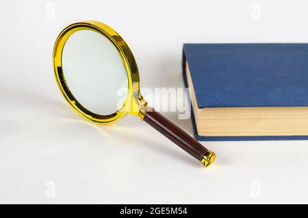 Loupe et livre fermé bleu en couverture rigide. Composition minimaliste sur fond blanc. Banque D'Images