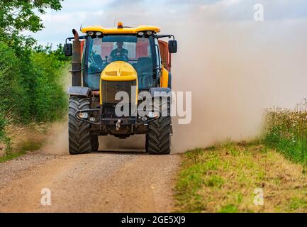 Un tracteur jaune tractant une remorque qui roule le long d'une étroite chenille de ferme carting du maïs au moment de la récolte et jette un nuage de poussière derrière lui Banque D'Images