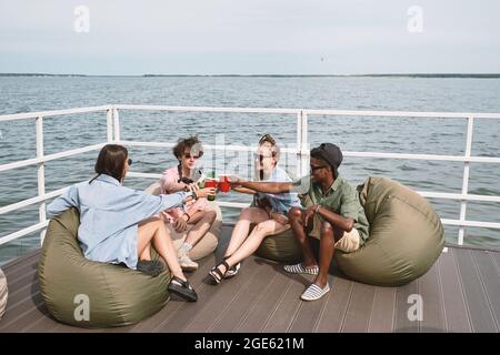 Groupe de jeunes ayant une petite fête sur la jetée en bois, ils sont assis dans des sacs de haricots et de toaster avec des boissons Banque D'Images