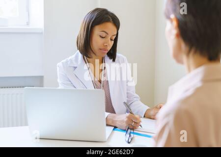 Femme médecin en uniforme médical écrivant les symptômes dans le presse-papiers pendant le rendez-vous avec le patient Banque D'Images