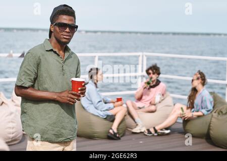 Jeune homme en casquette et lunettes de soleil tenant une tasse en plastique rouge, son ami se reposant sur des sacs de haricots en arrière-plan Banque D'Images