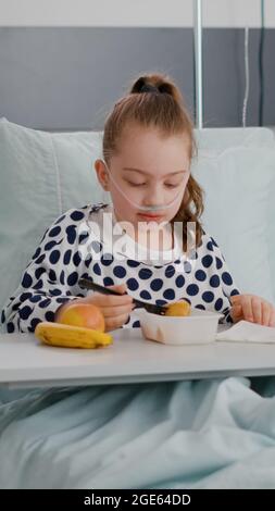 Portrait d'un petit enfant hospitalisé qui se repose au lit en mangeant un repas alimentaire sain pendant l'examen de récupération après une chirurgie médicale. Enfant malade portant un tube nasal à oxygène ayant une alimentation de petit déjeuner Banque D'Images