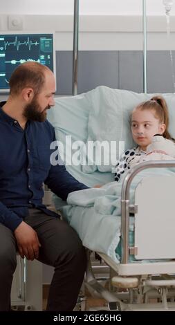 Une fille malade hospitalisée se reposant dans un lit portant un tube nasal à oxygène après avoir subi une chirurgie médicale pendant l'examen de la maladie dans la salle d'hôpital. Père expliquant le traitement de rétablissement Banque D'Images