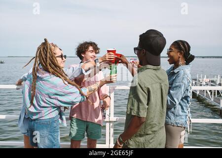 Joyeux jeunes gens joyeux qui toaster avec des verres et des bouteilles de bière lors d'une petite fête sur la jetée Banque D'Images