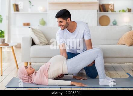 Un jeune mari arabe aide sa femme à faire de l'exercice de yoga, à s'entraîner sur un tapis à l'intérieur du salon. Épouses musulmanes aimantes s'exerçant ensemble pendant la pré Banque D'Images