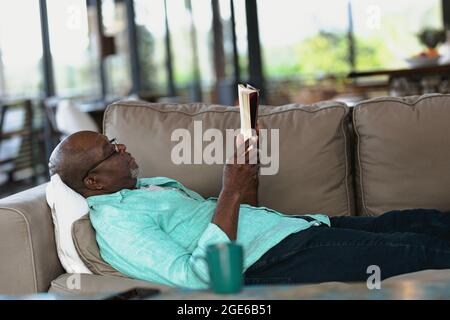 Relaxant homme afro-américain allongé sur le canapé et lisant un livre dans le salon moderne Banque D'Images