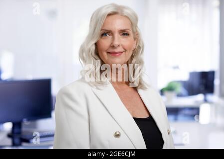 Portrait d'une femme d'affaires sénior aux cheveux gris debout au bureau tout en regardant l'appareil photo et en souriant. Banque D'Images