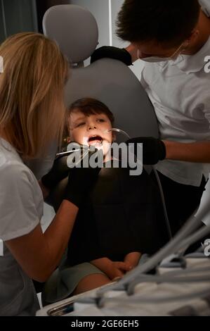 Instantané vertical d'une petite fille assise dans une chaise de dentiste et dentistes effectuant un examen des dents. Les médecins sont en uniforme et gants en latex. Fille portant un bavoir noir. Lumière sur la bouche des enfants Banque D'Images