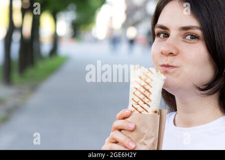 La femme Brunette surmange le shawarma dans une rue de la ville. Rouleau de pita de restauration rapide de rue avec de la viande et des légumes. Banque D'Images