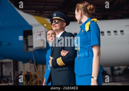 Pilote confiant en uniforme et aviateur lunettes de soleil marchant avec deux hôtesses de l'air en uniforme bleu devant le gros avion passager à l'intérieur Banque D'Images