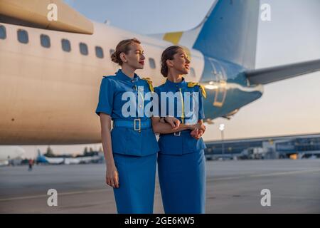 Deux belles hôtesses d'air en uniforme bleu souriant loin, debout devant un gros avion de passagers à l'aéroport au coucher du soleil Banque D'Images