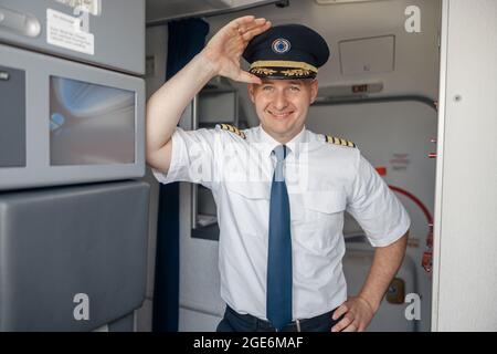 Joyeux pilote masculin en uniforme et chapeau souriant à la caméra tout en se tenant à l'intérieur de l'avion Banque D'Images