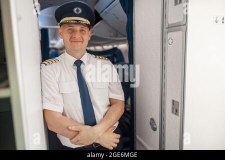 Joyeux pilote masculin en uniforme et chapeau en gardant les bras croisés et souriant à la caméra tout en se tenant à l'intérieur de l'avion Banque D'Images