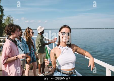 Portrait d'une jeune femme souriante dans des lunettes de soleil et crop top buvant de la bière à la fête extérieure sur la jetée Banque D'Images