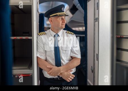 Un pilote homme attentif en uniforme et en chapeau, en gardant les bras croisés et en regardant de côté tout en se tenant à l'intérieur de l'avion Banque D'Images