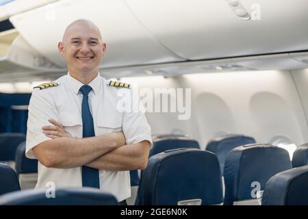Enjoué homme pilote en uniforme en gardant les bras croisés et souriant à la caméra tout en se tenant à l'intérieur de l'avion Banque D'Images