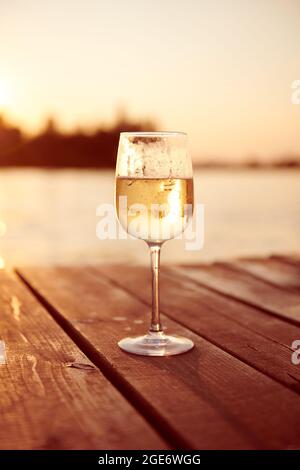 Vin blanc dans une coupe de champagne au coucher du soleil. Champagne froid mousseux sur une jetée en bois. Boisson individuelle avec lumière chaude. Photo de concept de détente. Banque D'Images