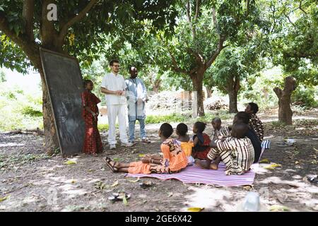 Un professeur africain présente un nouveau bénévole blanc à un petit groupe d'écoliers démunis dans une salle de classe en plein air dans un centre communautaire rural Banque D'Images