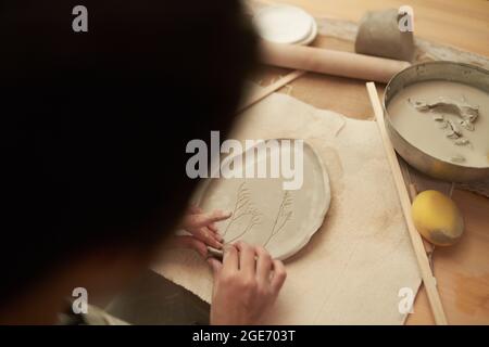 Vue arrière de la récolte artisanale anonyme travaillant dans la technique de construction à la main et de la plaque d'argile de façonnage avec motif floral à la table avec des outils en créati Banque D'Images