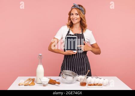 Portrait d'une belle jeune femme adulte tenant un ordinateur tablette et pointant vers lui écran blanc tout en se tenant près d'une table avec des ingrédients de boulangerie. IND Banque D'Images