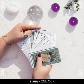 Les mains avec des clous et des anneaux violettes tiennent le pont de cartes de Tarot sur la surface blanche avec boule de cristal, bougies et pierres. Vue de dessus Banque D'Images