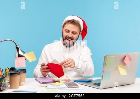 Homme à barbe enthousiaste dans un chapeau du père noël ouvrant une enveloppe rouge, allant lire la carte postale de vœux de vacances assis sur le lieu de travail avec un ordinateur portable. Studio intérieur s Banque D'Images