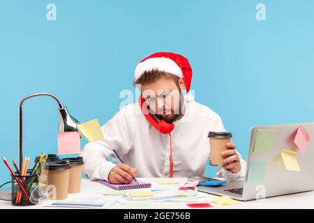 Homme au père noël chapeau assis sur le lieu de travail boire du café et parler téléphone fixe, écrire des informations dans le bloc-notes, travailler pendant les vacances Banque D'Images