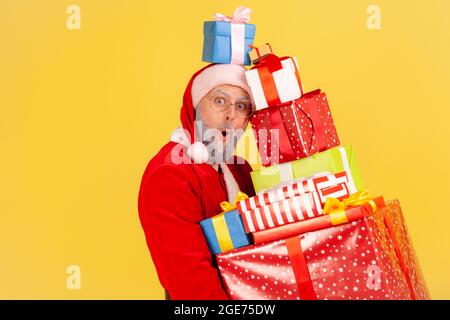 Homme âgé choqué avec une barbe en costume du père noël debout avec de nombreux cadeaux de Noël, regardant la caméra avec visage étonné, boîte de cadeaux lourds. Banque D'Images