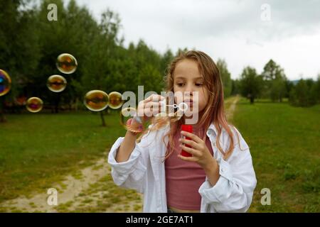 Jolie fille dans des vêtements décontractés soufflant des bulles de savon tout en se tenant sur la voie sur le jour couvert d'été dans la nature Banque D'Images