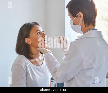 Femme d'âge moyen médecin généraliste en masque de protection du visage examinant la gorge du patient en clinique Banque D'Images