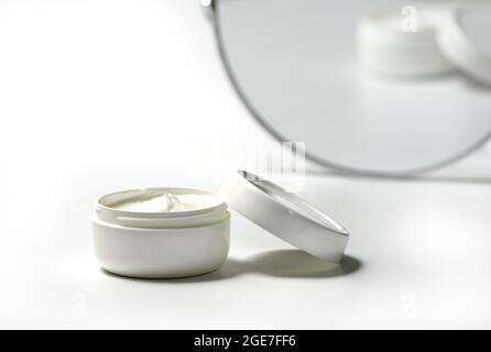 Pot blanc et casquette blanche avec crème pour le corps sur fond blanc avec miroir de maquillage dans le dos. Pot blanc avec crème pour le visage isolée. Produit cosmétique pour s Banque D'Images